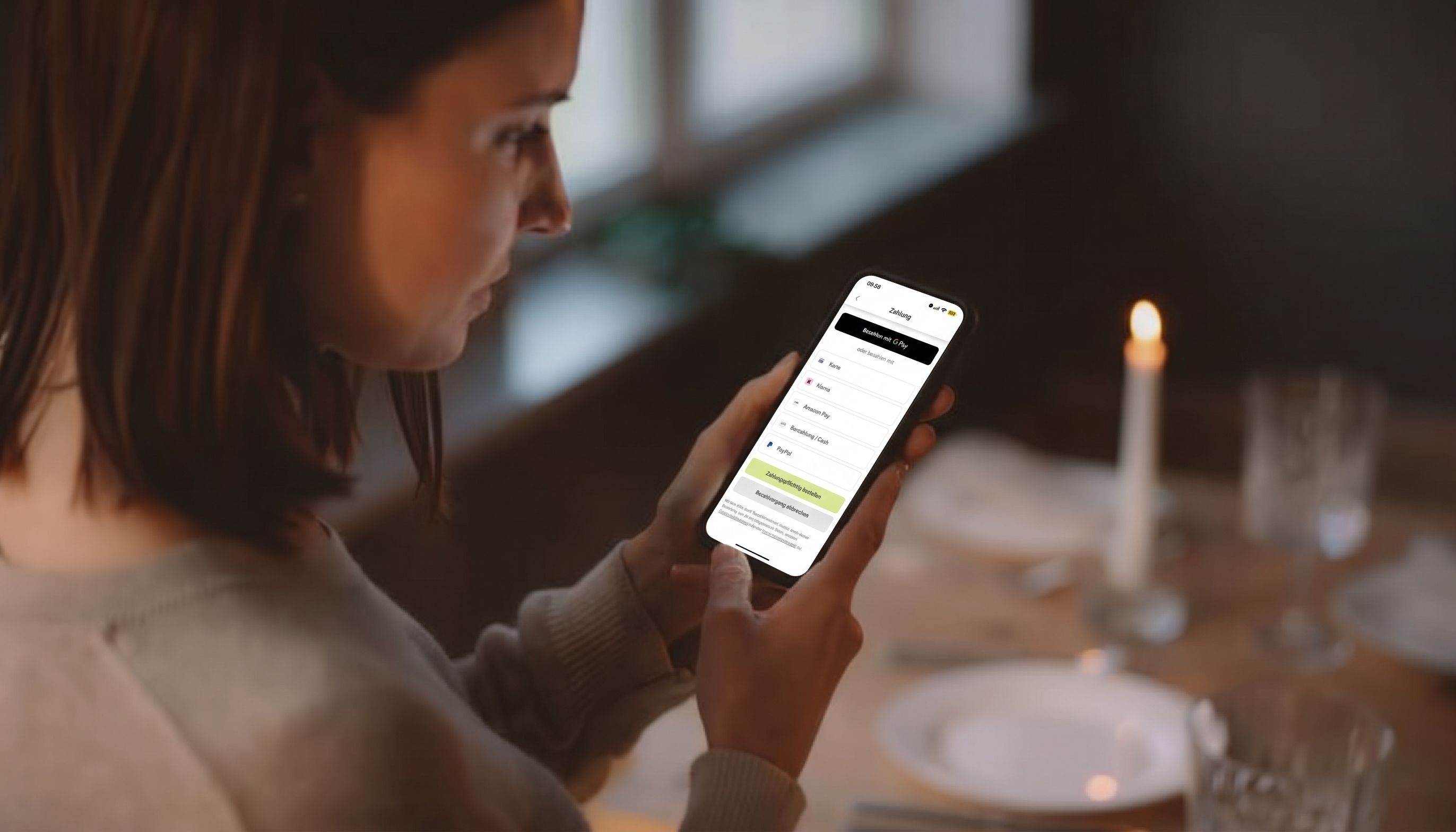 Eine Person zahlt in einer App, im Hintergrund ein Tisch mit einer Kerze.